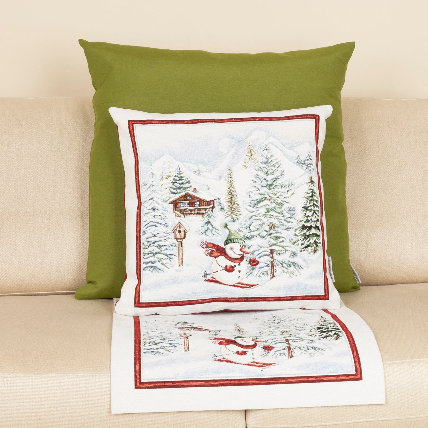 Quadratische Gobelin Chenille Kissenhülle und rechteckiger Tischläufer mit Motiv: Ein lustiger Schneemann mit rotem Schal fährt Ski in einer verschneiten Alpenlandschaft mit einer Berghütte. Platziert auf einem beige Sofa.
