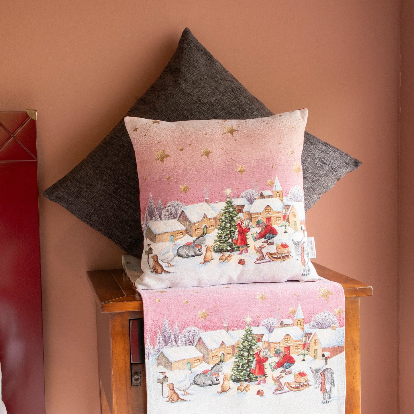 Quadratische Gobelin Kissenhülle und Tischläufer Detailreiche, fröhliche Weihnachtsdorf-Szene mit Kindern, Tieren und einem geschmückten Baum unter einem rosafarbenen Himmel mit Goldsternen platziert auf einer Holzkommode