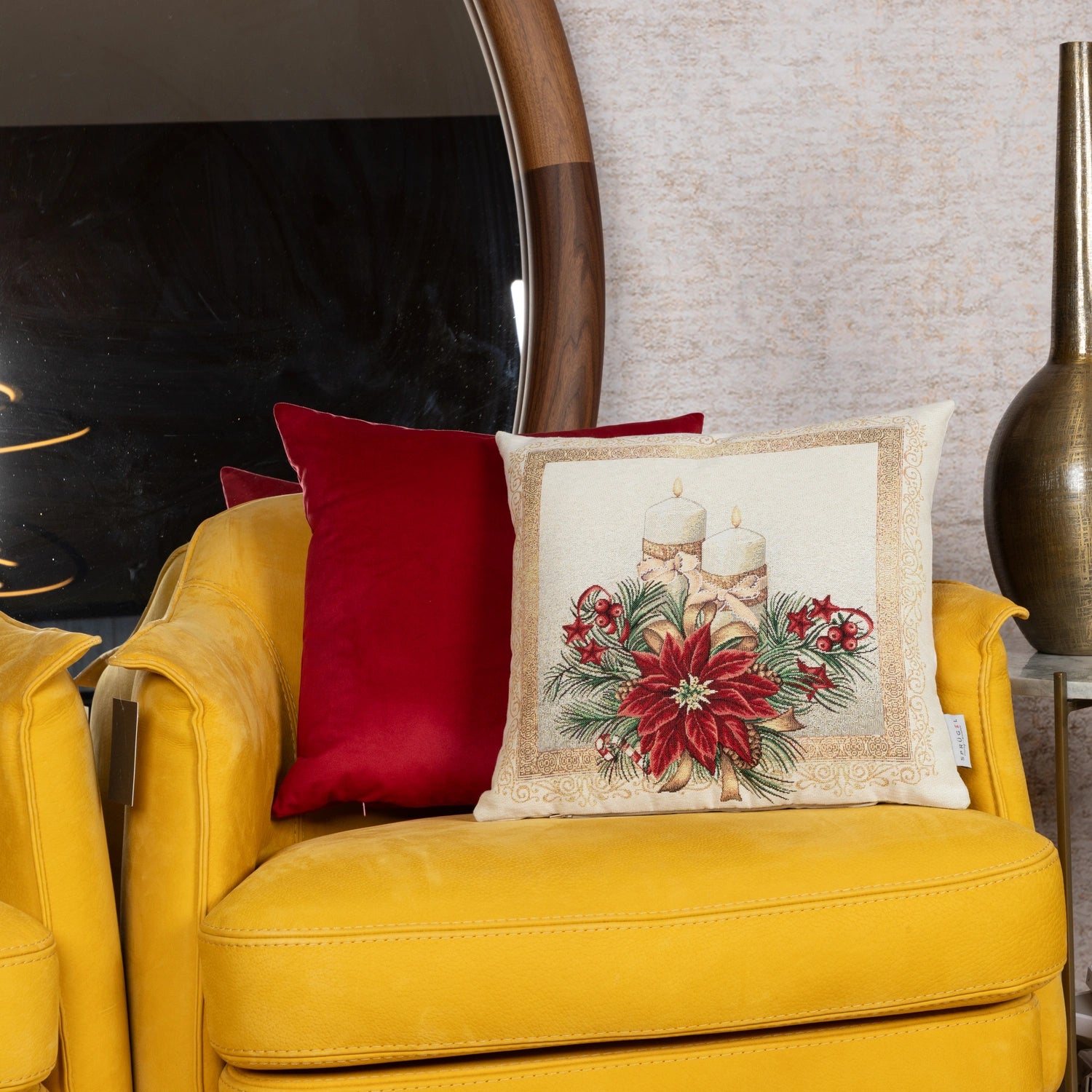 Quadratische Gobelin Kissenhülle mit weihnachtlichem Motiv: zwei helle Kerzen umgeben von roten Weihnachtssternen, Tannenzweigen und goldenen Ornamenten liegend auf einem gelben Sofa.